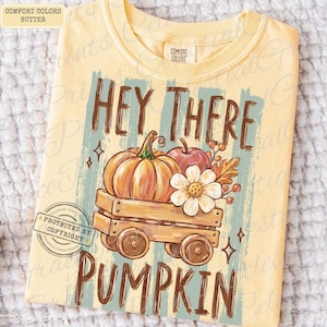 May include: A butter yellow t-shirt featuring a graphic print of a wooden wagon carrying a pumpkin, an apple, and a white flower. The text reads HEY THERE PUMPKIN in brown letters. The shirt has a Comfort Colors label at the inner neckline.