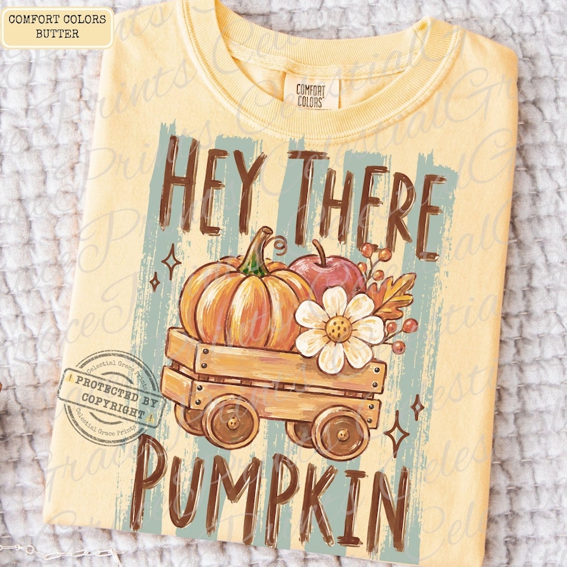 May include: A butter yellow t-shirt featuring a graphic print of a wooden wagon carrying a pumpkin, an apple, and a white flower. The text reads HEY THERE PUMPKIN in brown letters. The shirt has a Comfort Colors label at the inner neckline.