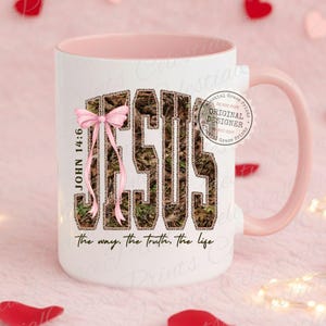 May include: A white ceramic mug with a pink interior and handle. The mug features the word "JESUS" in a camouflage pattern with a pink bow. The text "JOHN 14:6" and "the way, the truth, the life" are also visible.