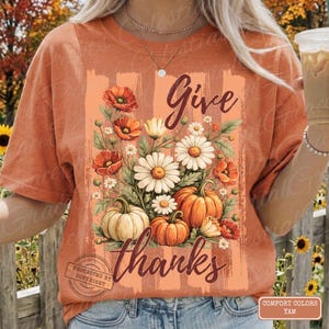 Può includere: T-shirt arancione con grafica floreale e zucche. Il design include le parole "Give thanks" in un font corsivo, insieme a illustrazioni di zucche, margherite e papaveri. La maglietta è del marchio Comfort Colors.