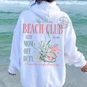 Puede incluir: Sudadera con capucha blanca con un dise&ntilde;o de tem&aacute;tica playera. El dise&ntilde;o incluye el texto "Beach Club", "Mom Off Duty" y "Salty Hair Ocean Air". Tambi&eacute;n se muestra una estrella de mar, una concha y algas marinas.