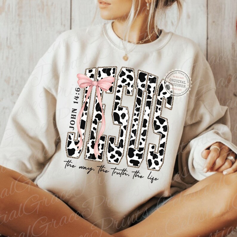 Puede incluir: Sudadera color crema con la palabra "JESUS" en un dise&ntilde;o de estampado de vaca en blanco y negro. Un lazo rosa y el texto "JOHN 14:6" est&aacute;n en el lado izquierdo. La frase "the way, the truth, the life" est&aacute; impresa debajo.