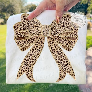 Puede incluir: Una camiseta blanca con un gran diseño de lazo con estampado de leopardo. El lazo tiene un contorno dorado y un patrón texturizado. El texto "Celestial Grace Prints" es visible en el fondo.