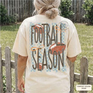 May include: An ivory t-shirt featuring a back graphic with the text FOOTBALL SEASON in large dark brown letters. The design includes a light blue rectangular background with yellow polka dots, three brown football illustrations, and orange bows.