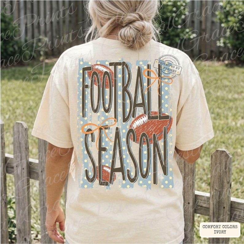 May include: An ivory t-shirt featuring a back graphic with the text FOOTBALL SEASON in large dark brown letters. The design includes a light blue rectangular background with yellow polka dots, three brown football illustrations, and orange bows.