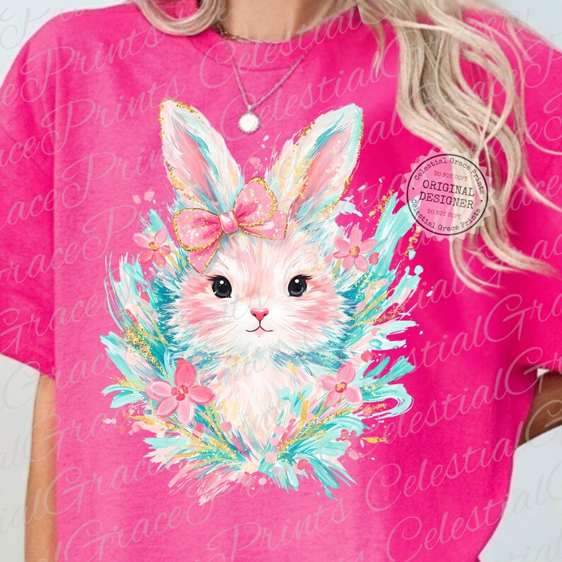 May include: Pink t-shirt featuring a whimsical bunny design. The bunny has a pink bow, surrounded by blue and gold accents, and pink flowers. The text "Celestial Grace Prints" is visible on the shirt.