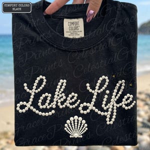 May include: Black t-shirt featuring the words "Lake Life" in white pearl embellishments, with a seashell design. Made from Comfort Colors cotton.