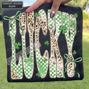 May include: Black t-shirt featuring the word "LUCKY" in a dripping style. The letters are decorated with a green and white checked pattern, leopard print, and shamrocks. Green bows adorn the top and bottom. The text "COMFORT COLORS BLACK" is in the top left corner.