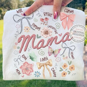 May include: A white t-shirt with the word "Mama" in pink script, surrounded by floral and butterfly illustrations. The design includes words like "Wise," "Brave," and "Loved," with bow accents. The shirt is held up by a hand.