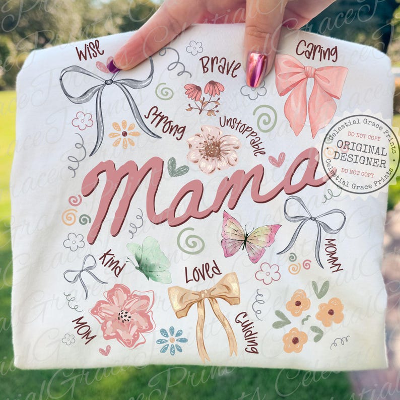 May include: A white t-shirt with the word "Mama" in pink script, surrounded by floral and butterfly illustrations. The design includes words like "Wise," "Brave," and "Loved," with bow accents. The shirt is held up by a hand.