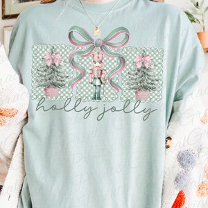 May include: A pale blue t-shirt with a Christmas-themed graphic. The design includes a nutcracker, Christmas trees, and a large bow in green and pink. The words "holly jolly" are written in a script font below the design.