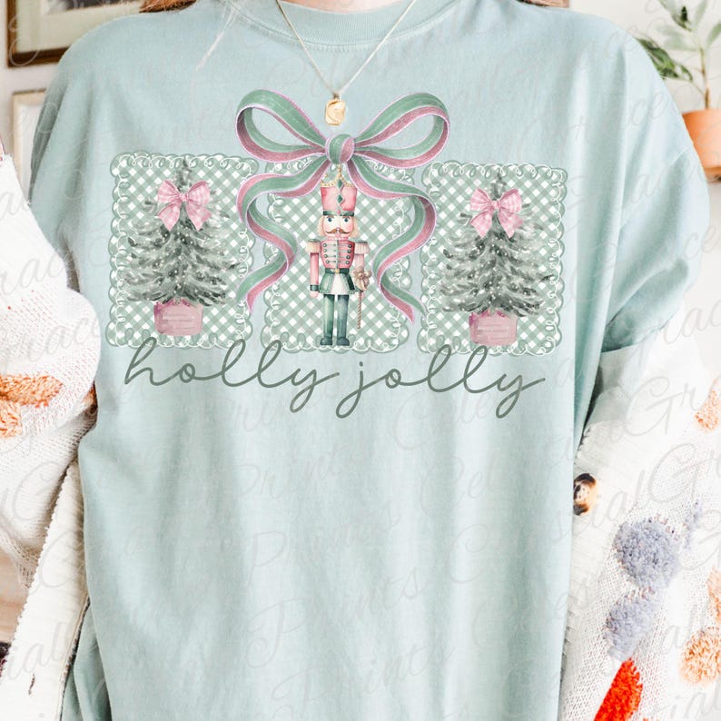 May include: A pale blue t-shirt with a Christmas-themed graphic. The design includes a nutcracker, Christmas trees, and a large bow in green and pink. The words "holly jolly" are written in a script font below the design.
