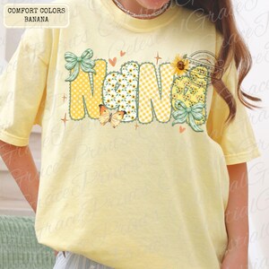 May include: A light yellow t-shirt with the word "Nana" in a playful design. The letters feature polka dots, gingham, and floral patterns, with butterflies, bows, and hearts. The shirt is labeled "Comfort Colors Banana".