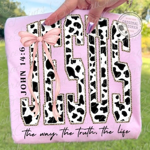 Puede incluir: Una sudadera rosa claro con la palabra "JESUS" en un dise&ntilde;o de estampado de vaca con un contorno de estampado de leopardo. El texto "JOHN 14:6" est&aacute; a la izquierda, y la frase "the way, the truth, the life" est&aacute; en la parte inferior. Un lazo rosa est&aacute; a la izquierda.