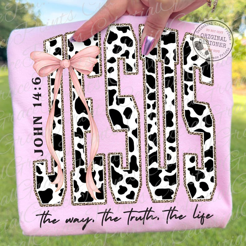 Puede incluir: Una sudadera rosa claro con la palabra "JESUS" en un dise&ntilde;o de estampado de vaca con un contorno de estampado de leopardo. El texto "JOHN 14:6" est&aacute; a la izquierda, y la frase "the way, the truth, the life" est&aacute; en la parte inferior. Un lazo rosa est&aacute; a la izquierda.