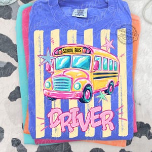 Può includere: Una t-shirt blu con la grafica di uno scuolabus giallo con la scritta SCHOOL BUS sopra e la parola DRIVER in grandi lettere rosa sotto. Il design include strisce verticali giallo chiaro e diverse forme a stella sullo sfondo.