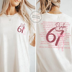 May include: Cream-colored t-shirt featuring the text "Psalm 67" in a burgundy font with a pink bow. The back of the shirt displays the full text of Psalm 67 in a burgundy font. A casual, comfortable tee.