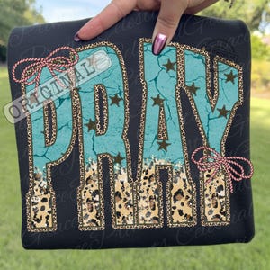 May include: Black sweatshirt featuring the word "PRAY" in turquoise, leopard print, and brown star accents. The word "ORIGINAL" is printed on the upper left corner. Pink bows are on the upper left and right sides.