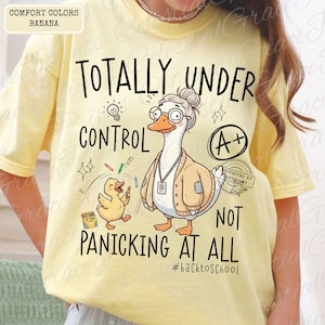 May include: Yellow Comfort Colors t-shirt with the text "TOTALLY UNDER CONTROL NOT PANICKING AT ALL" and a cartoon goose and duck graphic. The shirt has a crew neck and short sleeves.
