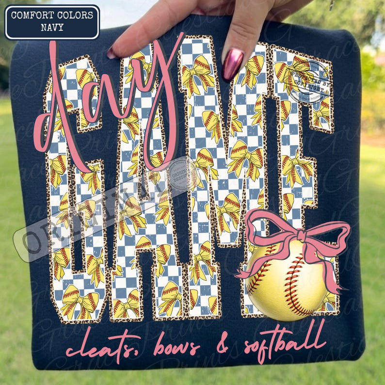 May include: Navy blue sweatshirt with the words "Day Game" in pink script and block letters. The letters are filled with a blue and white checkered pattern and yellow bows. A softball with a pink bow is on the right. The words "cleats, bows & softball" are at the bottom.