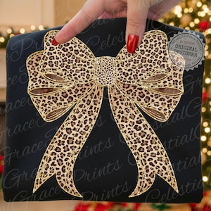 May include: Black sweatshirt featuring a large leopard print bow with gold glitter trim. The bow design includes a detailed leopard pattern and is centered on the front of the shirt. The text "Celestial Grace Prints" is visible.