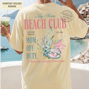Puede incluir: Camiseta de color pl&aacute;tano con el texto "Salty Moms Beach Club" y un gr&aacute;fico de concha. El dise&ntilde;o incluye una estrella de mar, una perla y las palabras "Mom Off Duty". La camiseta tambi&eacute;n tiene el texto "Powered by iced coffee & beach days."