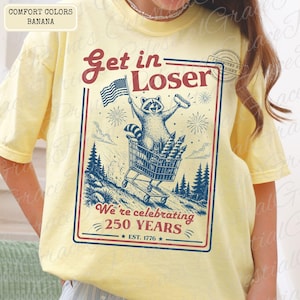 May include: Yellow t-shirt with a graphic featuring a raccoon in a shopping cart, holding an American flag and a hot dog. The text reads "Get in Loser" and "We're celebrating 250 YEARS EST. 1776."
