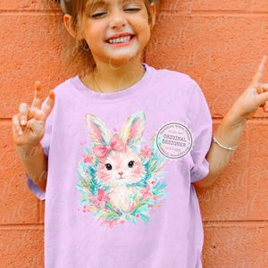 May include: A light purple t-shirt featuring a cute bunny illustration with a pink bow, surrounded by flowers and leaves. The bunny has a pink nose and is surrounded by a wreath of flowers. The text "ORIGINAL DESIGNER" is printed on the shirt.