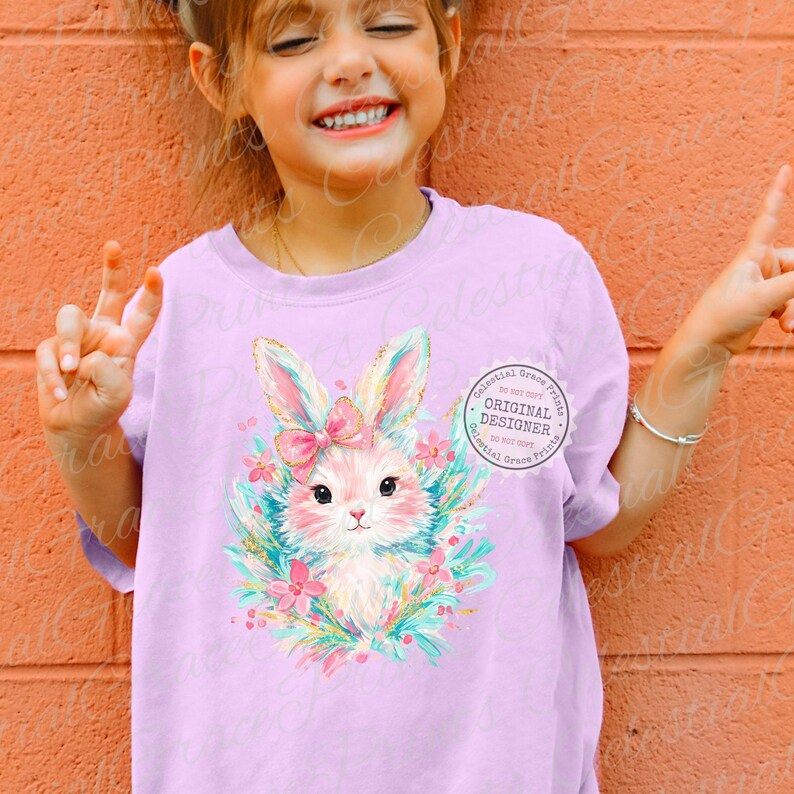 May include: A light purple t-shirt featuring a cute bunny illustration with a pink bow, surrounded by flowers and leaves. The bunny has a pink nose and is surrounded by a wreath of flowers. The text "ORIGINAL DESIGNER" is printed on the shirt.
