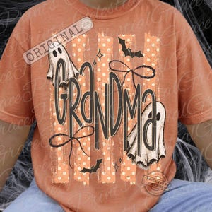 May include: A burnt orange t-shirt featuring a graphic print with the word GRANDMA in large black letters. The design includes two white ghosts, black bats, and thin black line-art bows over a background of vertical beige stripes with white dots.