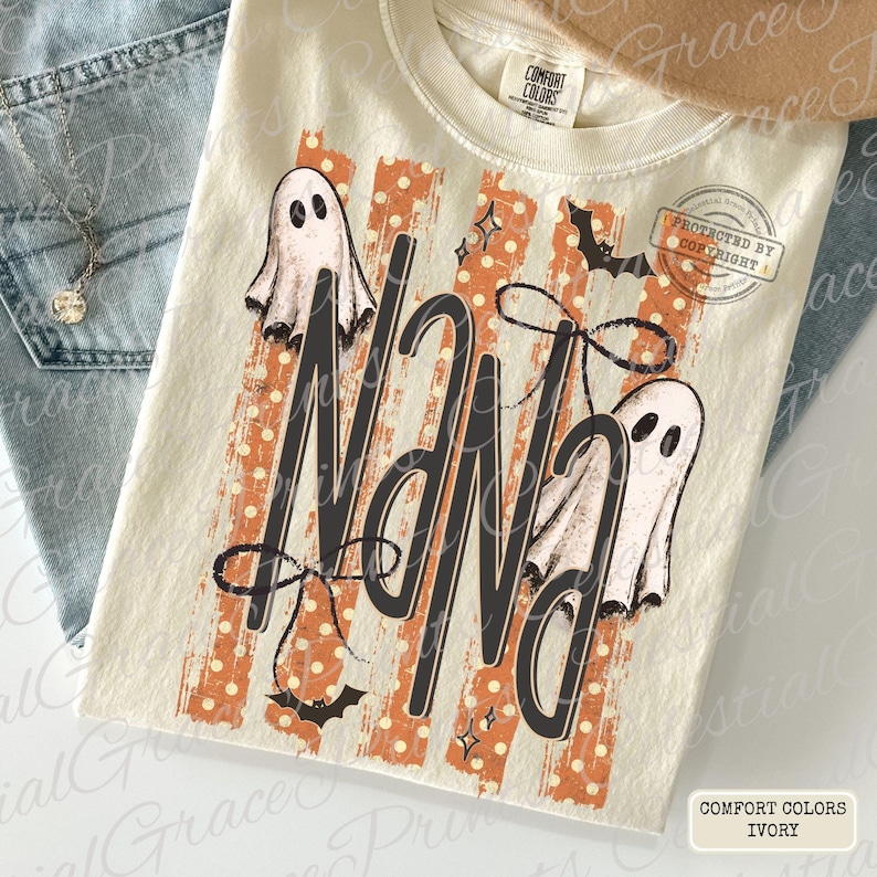 May include: An ivory t-shirt with a graphic print featuring the word Nana in large black letters. The design includes two white ghosts, black bows, small stars, and bats over a background of vertical orange and white polka dot stripes.
