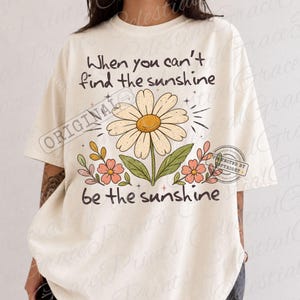 May include: An off-white short-sleeve t-shirt featuring a large daisy graphic in the center with smaller pink flowers and green leaves. The text reads, "When you can't find the sunshine be the sunshine," printed in a dark brown, rounded font.