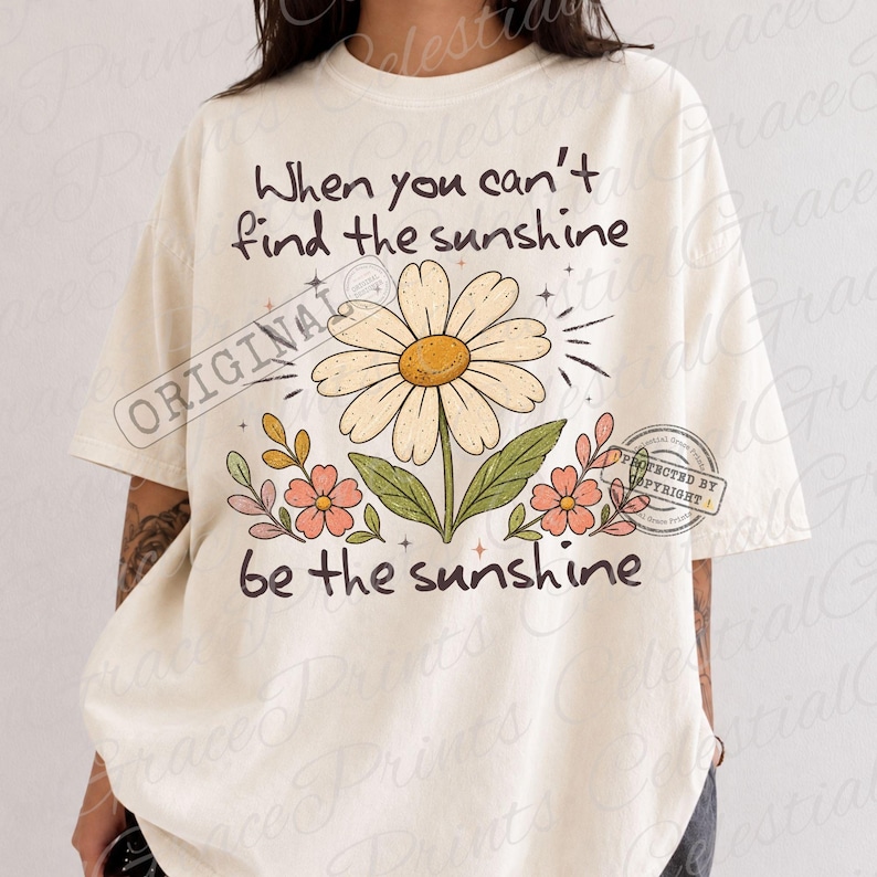 May include: An off-white short-sleeve t-shirt featuring a large daisy graphic in the center with smaller pink flowers and green leaves. The text reads, "When you can't find the sunshine be the sunshine," printed in a dark brown, rounded font.