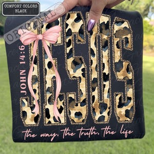 May include: Black sweatshirt featuring the word "JESUS" in a leopard print pattern. A pink bow and the text "JOHN 14:6" are on the left. The phrase "the way, the truth, the life" is printed at the bottom of the garment.