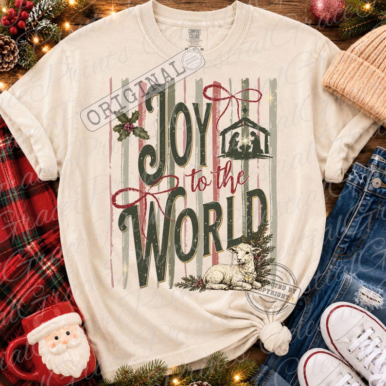 Puede incluir: Una camiseta de manga corta color crema con el texto "JOY to the WORLD" en verde oscuro y rojo. El dise&ntilde;o incluye una silueta de escena de natividad, un peque&ntilde;o cordero, hojas de acebo y rayas verticales en tonos apagados de rojo, verde y crema.