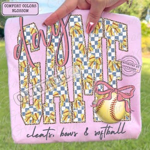 May include: Pink sweatshirt with the words "Day Game" in a pink script font. The letters are filled with a blue and white checkered pattern, yellow bows, and softball details. A softball with a pink bow is on the bottom right. The text "cleats, bows & softball" is at the bottom.
