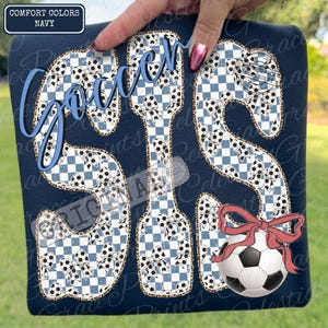 May include: Navy blue t-shirt with the word "Soccer" in blue script. The letters "SIS" are filled with a blue and white checkered pattern and soccer ball accents. A soccer ball with a red bow is on the bottom right.