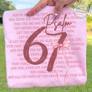 May include: A light pink t-shirt featuring the text of Psalm 67 in a dark red font. A large number 67 is centered on the shirt, with a pink bow and ribbon. The shirt also has the text "Celestial Grace Prints" and "Original Designer".