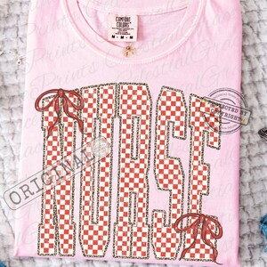 Peut inclure: T-shirt rose clair avec le mot "MAGIC" en motif &agrave; carreaux rouges et blancs, bord&eacute; d'un motif l&eacute;opard. Le t-shirt comporte deux n&oelig;uds rouges et une &eacute;tiquette Comfort Colors.