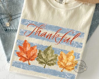Könnte beinhalten: Elfenbeinfarbenes Comfort Colors T-Shirt mit dem Wort "Thankful" in roter Schrift über blauen Streifen. Darunter befinden sich drei bunte Herbstblätter in Orange, Grün und Gelb.