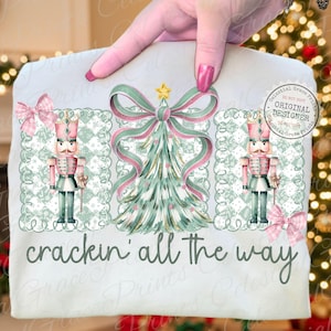 以下が含まれることがあります： クリスマスデザインの明るい色のTシャツ。デザインには、リボン付きのクリスマスツリー、2つのくるみ割り人形、「crackin' all the way」というフレーズが含まれています。デザインはピンク、グリーン、ゴールドの色合いを使用しています。