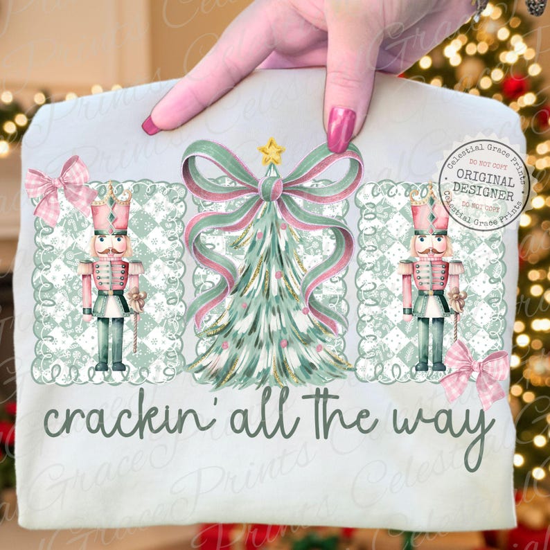 以下が含まれることがあります： クリスマスデザインの明るい色のTシャツ。デザインには、リボン付きのクリスマスツリー、2つのくるみ割り人形、「crackin' all the way」というフレーズが含まれています。デザインはピンク、グリーン、ゴールドの色合いを使用しています。