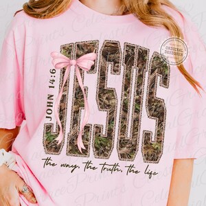 May include: Pink t-shirt with the word "JESUS" in a camouflage and leopard print design. A pink bow and the text "JOHN 14:6" are on the left. The phrase "the way, the truth, the life" is printed below.