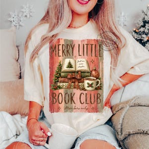 May include: A cream-colored t-shirt featuring a graphic of a stack of books, a Christmas tree, and a mug with holly. The text reads MERRY LITTLE BOOK CLUB, Just one more chapter, and Members only. The design has a rustic, wood-plank background.