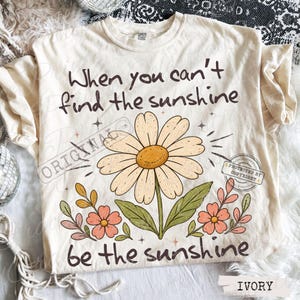 May include: An ivory t-shirt featuring a large daisy illustration in the center with smaller flowers on the sides. The text reads, "When you can't find the sunshine, be the sunshine." The design includes small star accents and a brown color palette.