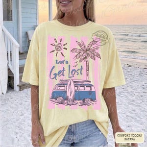 May include: A yellow Comfort Colors t-shirt with a graphic design featuring a sun, palm tree, and a blue van with a surfboard. The text "Let's Get Lost" is displayed in blue script. The shirt is a banana color.