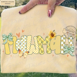 May include: A light yellow t-shirt with the word "MAMA" in large, colorful letters. The letters feature patterns like sunflowers, gingham, and chevron. The design includes bows, hearts, and a butterfly. The shirt is a comfort color.