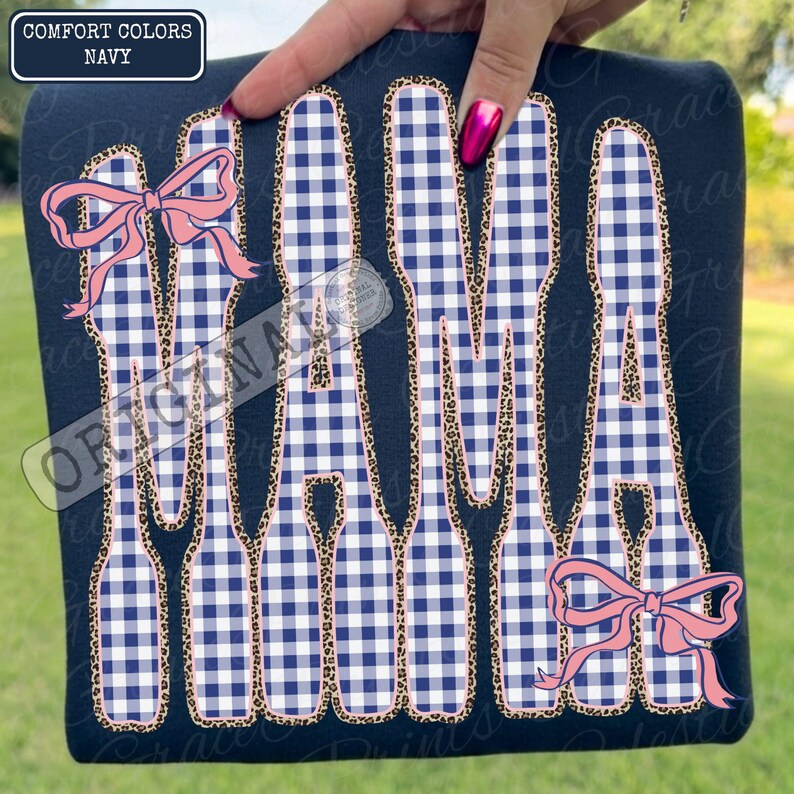 May include: Navy blue t-shirt featuring the word "MAMA" in a white and blue gingham design, edged with a leopard print. Pink bows adorn the top left and bottom right. A label in the top left corner reads "COMFORT COLORS NAVY".