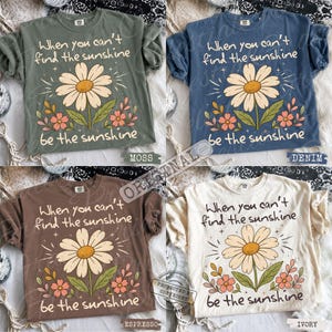 May include: Four short-sleeve t-shirts in moss green, denim blue, espresso brown, and ivory. Each shirt features a central graphic of a large daisy with smaller pink flowers and the text: When you can't find the sunshine be the sunshine.