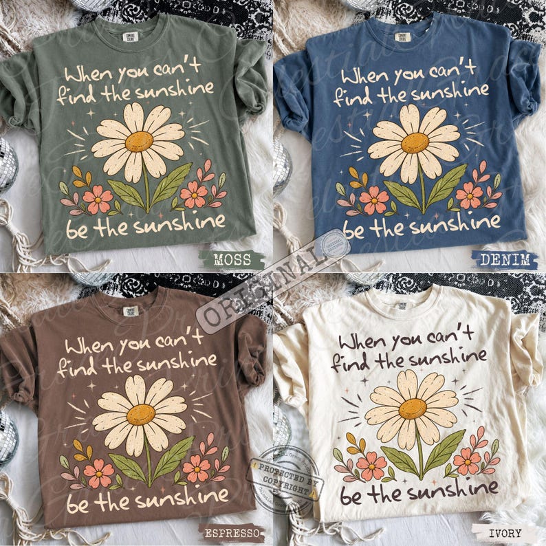 May include: Four short-sleeve t-shirts in moss green, denim blue, espresso brown, and ivory. Each shirt features a central graphic of a large daisy with smaller pink flowers and the text: When you can't find the sunshine be the sunshine.